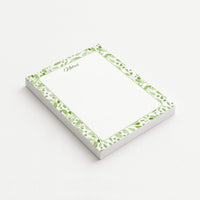 Sage Botanica memo pad shown at an angle with a watercolor border of green leaves, berries, and botanical sprigs surrounding a subtly dotted writing area.