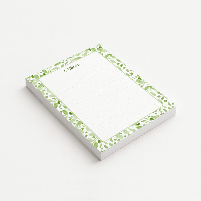 Sage Botanica memo pad shown at an angle with a watercolor border of green leaves, berries, and botanical sprigs surrounding a subtly dotted writing area.
