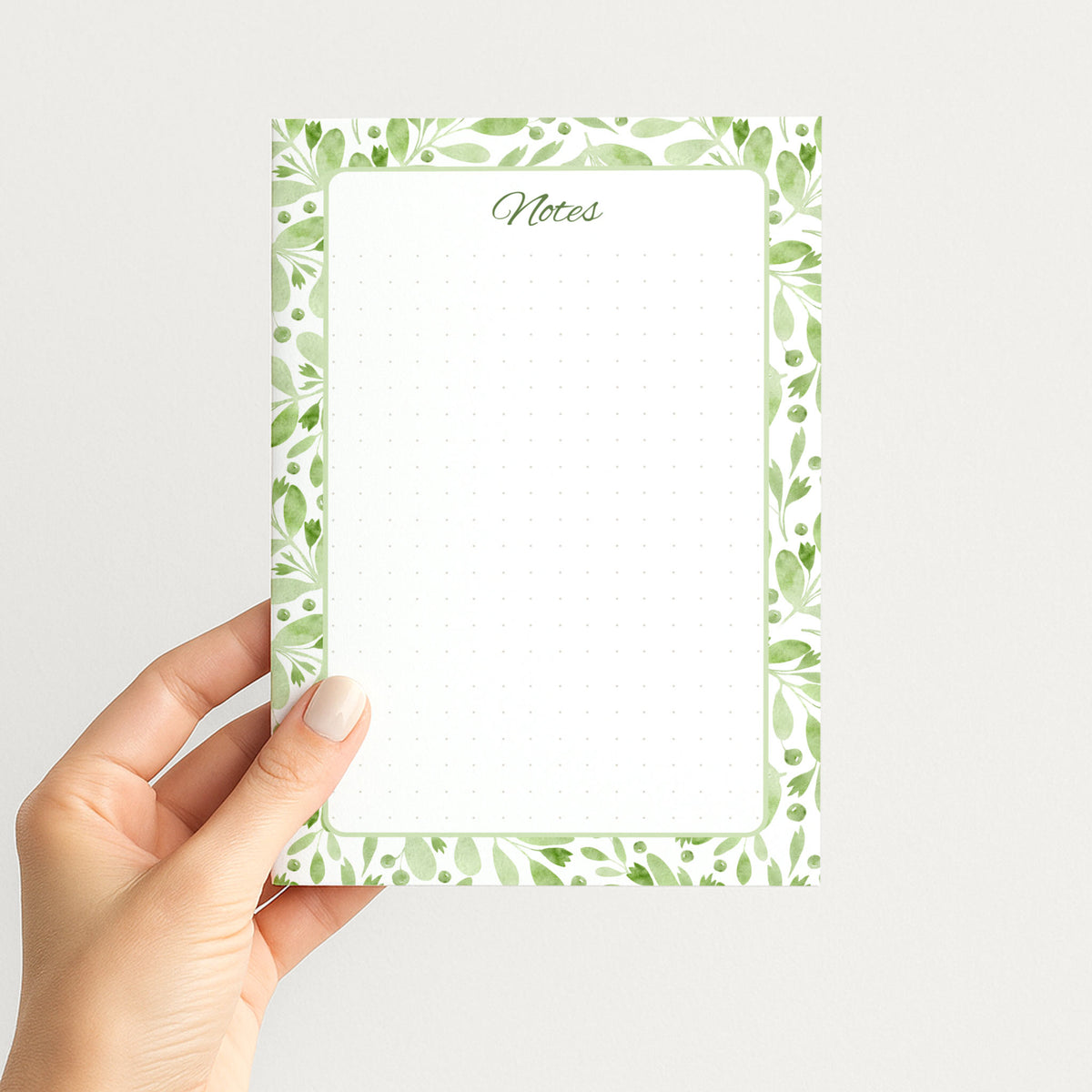 Hand holding the Sage Botanica memo pad upright, showing its soft green botanical border, script Notes heading, and dotted writing field.