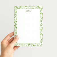 Hand holding the Sage Botanica memo pad upright, showing its soft green botanical border, script Notes heading, and dotted writing field.