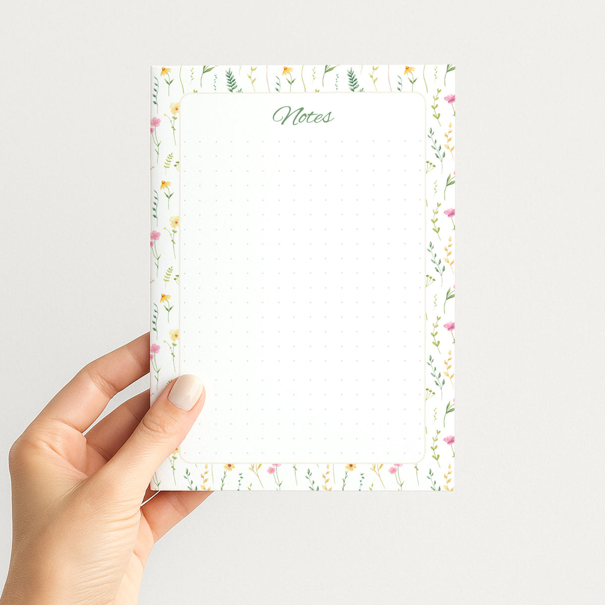 Hand holding the Wildflower Meadow memo pad upright, showing its airy watercolor floral border, green script Notes heading, and dotted writing field.