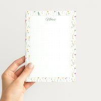 Hand holding the Wildflower Meadow memo pad upright, showing its airy watercolor floral border, green script Notes heading, and dotted writing field.