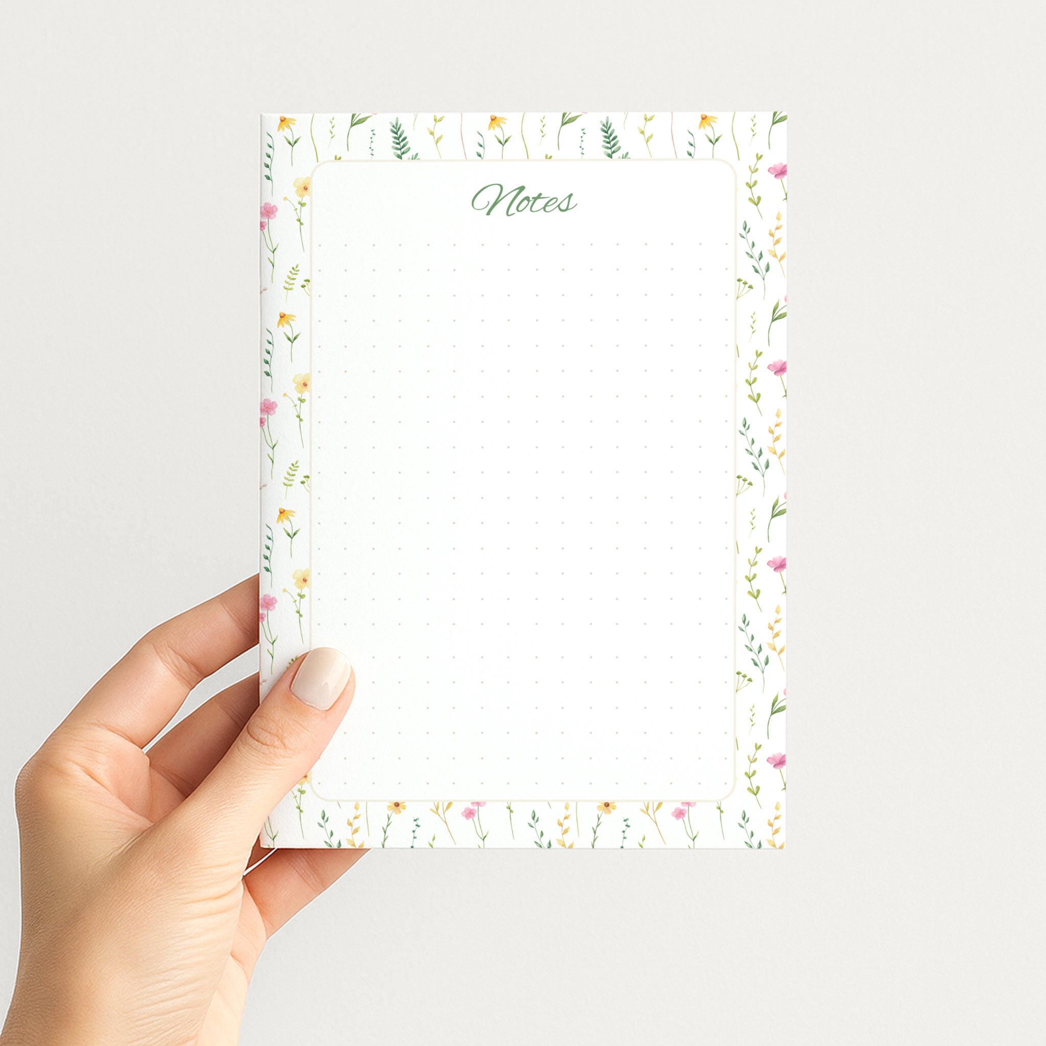 Hand holding the Wildflower Meadow memo pad upright, showing its airy watercolor floral border, green script Notes heading, and dotted writing field.
