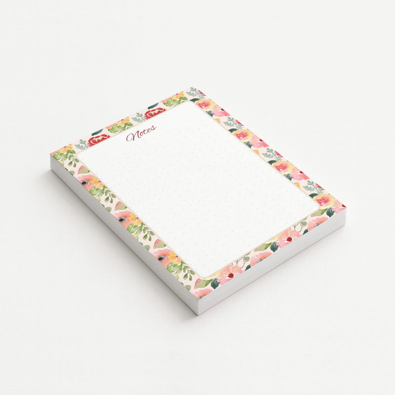 Painter’s Garden memo pad shown at an angle with a watercolor border of blush, coral, yellow, and red flowers surrounding a subtly dotted writing area.