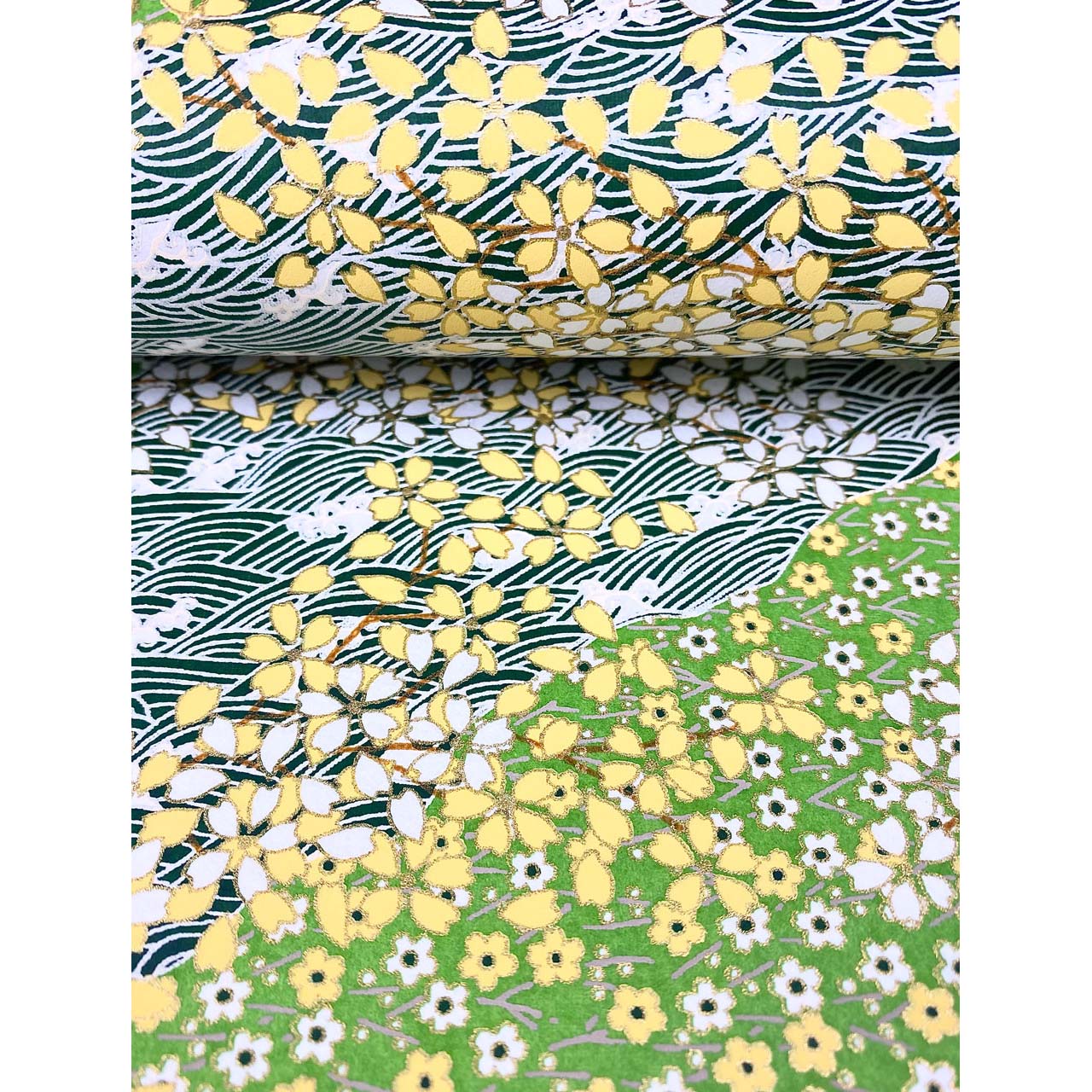 Close-up of hand silk-screen printed Chiyogami paper showing green garden bands, delicate blossoms, leafy branches, and dark wave patterns. This paper is bent like the page of a book.