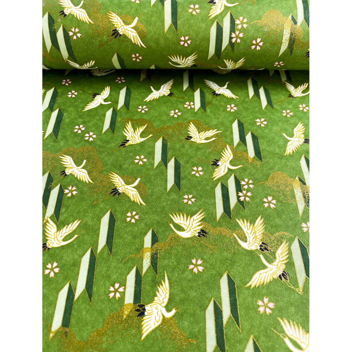 Close-up of hand silk-screen printed Chiyogami paper showing ivory cranes in flight, small blossoms, and angular motifs on green. Paper is bent like the page of a book.