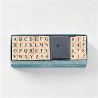 A wooden alphabet and number block set in a teal box, with a black square eraser in the center, photographed from above on a white background.