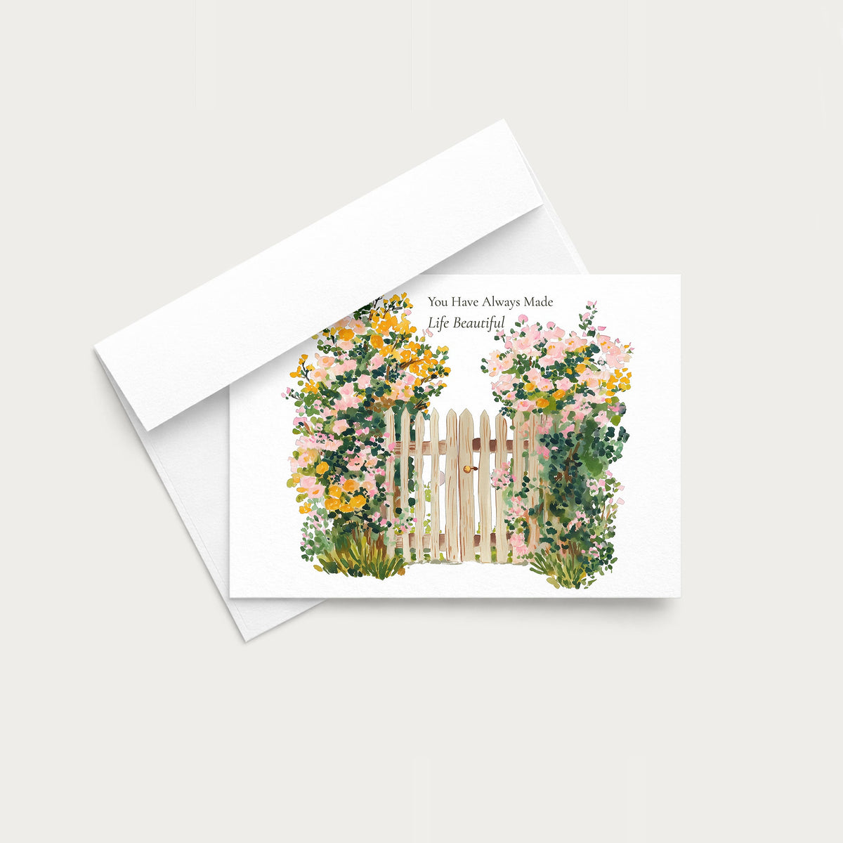 A white greeting card with a watercolor illustration of pink, yellow, and green flowers around a wooden fence, shown with a white envelope on a light gray background.
