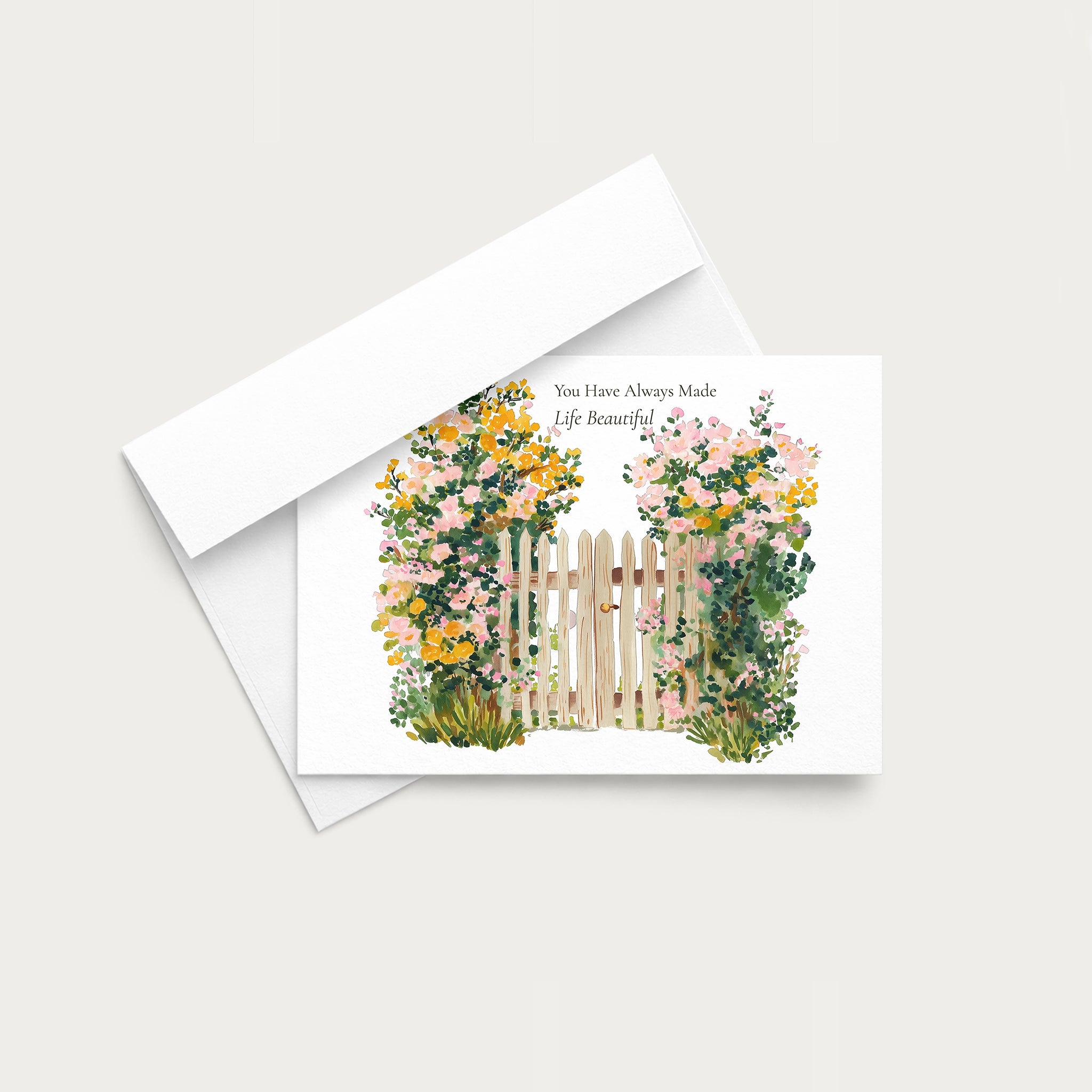 A white greeting card with a watercolor illustration of pink, yellow, and green flowers around a wooden fence, shown with a white envelope on a light gray background.